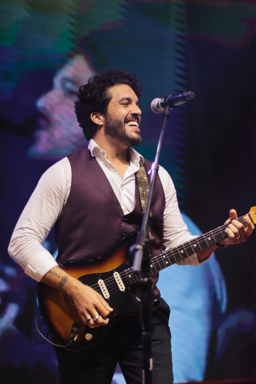 Bruno sorrindo ao microfone com guitarra, telão colorido ao fundo