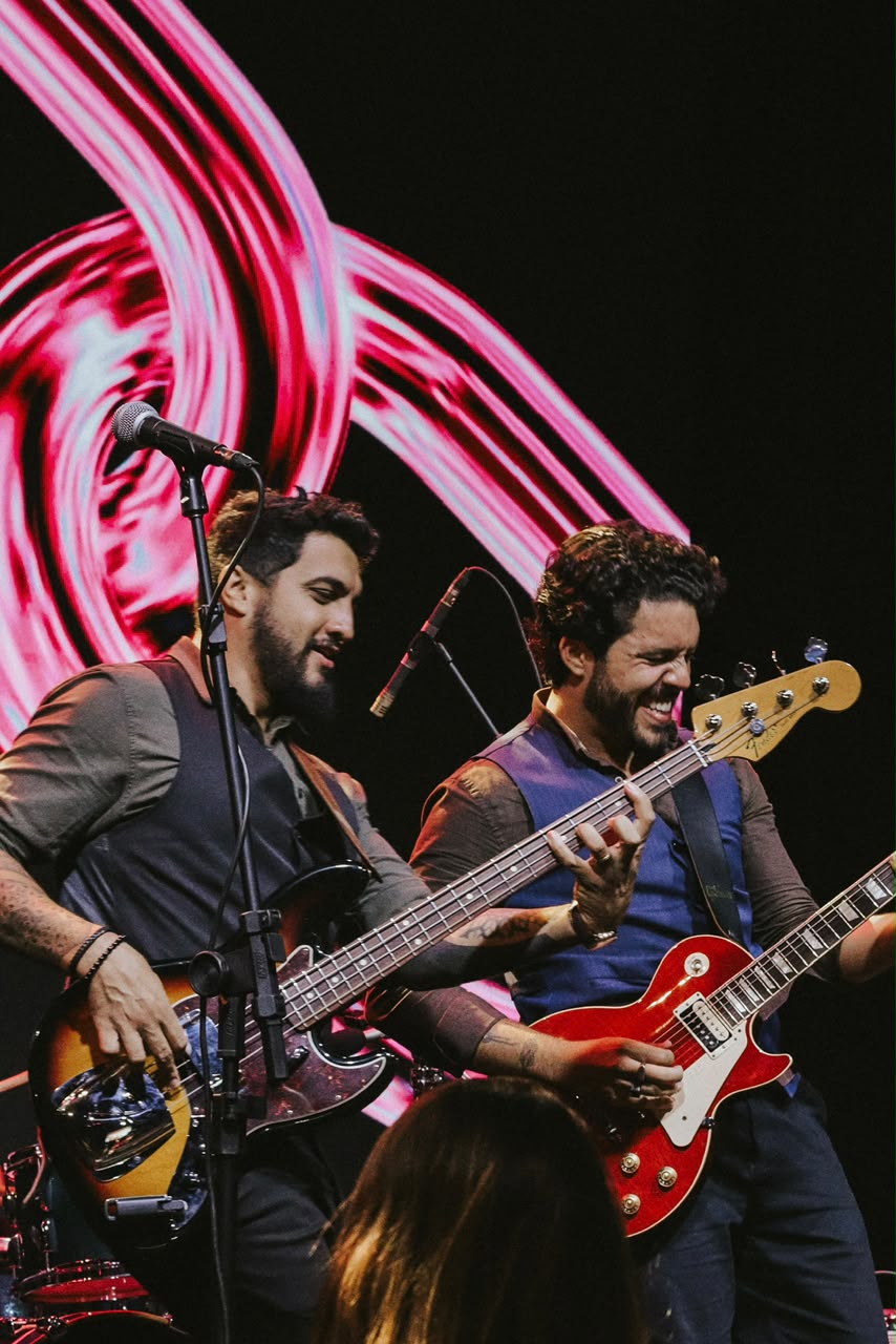 Rapha e Bruno rindo juntos no palco, com telão rosa ao fundo
