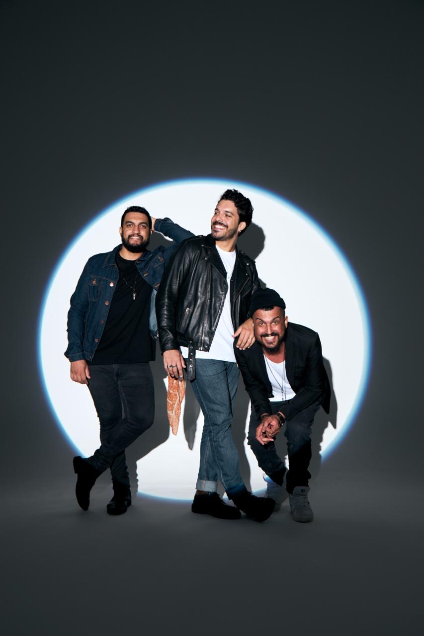 Os três integrantes sorrindo dentro de um círculo de luz